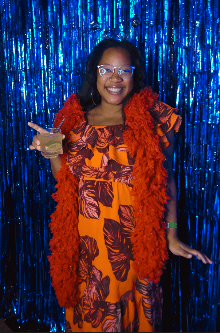 A guest in an orange floral dress and red boa in front of a blue tinsel backdrop