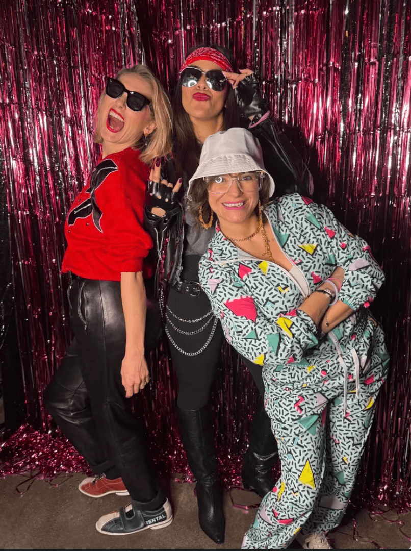 An 80s-themed party trio posing in front of a pink tinsel backdrop