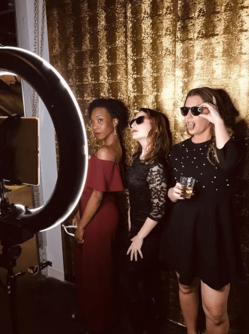 Three women in sunglasses posing in front of a gold sequin backdrop